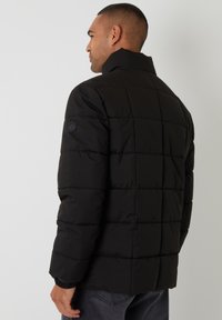 Schwarze Puffy-Jacke mit quadratischem Steppmuster, hohem Kragen, elastischen Bündchen und einem Logo-Patch am linken Ärmel.