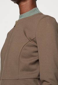 Gros plan sur une personne portant une veste zippée taupe par-dessus un haut à col montant côtelé léger vert, montrant le haut du torse et le cou.