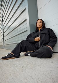 Svart fleecejacka med två fickor, parat med svarta vida byxor och svarta sneakers med bruna sulor och vita snören. Slät bakgrund.