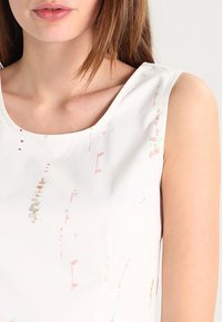 Haut sans manches blanc avec des motifs multicolores subtils. Le tissu a une texture lisse avec un col rond et des coutures minimales.