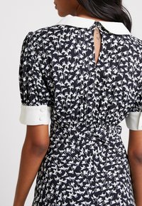 Robe noire avec motifs floraux blancs, manches courtes bouffantes et col blanc. Décorée d'une ouverture en forme de trou de serrure dans le dos et d'une taille froncée.