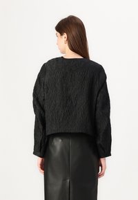 Black cropped jacket with a textured, wrinkled fabric. Features long sleeves and a wide neckline, paired with a black leather skirt.