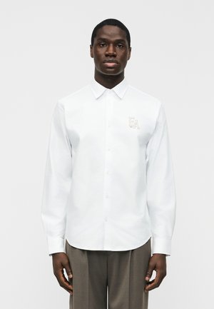 Uomo che indossa una camicia bianca a maniche lunghe con bottoni e logo "EA" sul petto a sinistra, e pantaloni marroni, in piedi davanti a uno sfondo semplice.