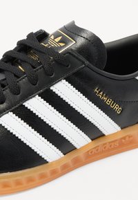 Sneaker Adidas Hamburg noire avec des rayures blanches, semelle en caoutchouc, lacets noirs et détails en or sur la languette et le côté.