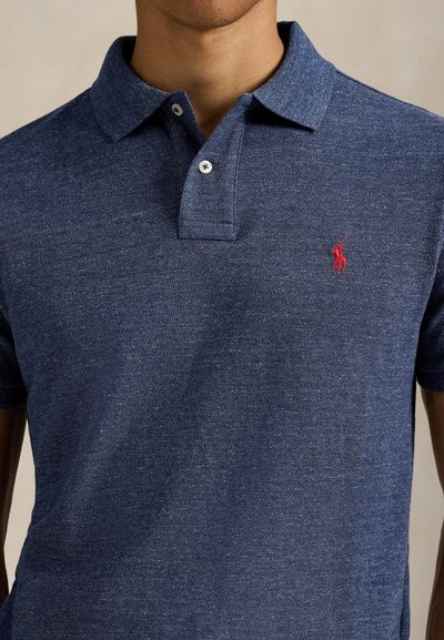 Homme portant un polo bleu à manches courtes avec un logo de cheval rouge brodé sur le côté gauche de la poitrine.
