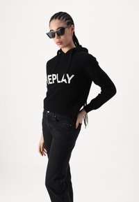 Melna ribota kapuce ar baltu "REPLAY" logo, pārī ar melniem džinsiem. Modelis valkā melnas taisnstūrveida saulesbrilles un ir ar pītām matu frizūrām.