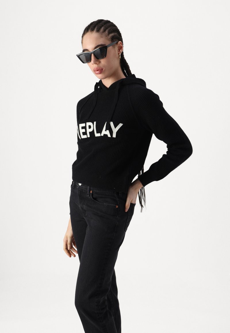 Schwarze Rippstrick-Hoodie mit weißem "REPLAY"-Logo, kombiniert mit schwarzen Jeans. Das Model trägt schwarze rechteckige Sonnenbrillen und hat geflochtenes Haar.