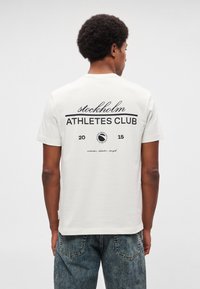 Biele bavlnené tričko s čiernym textom „stockholm ATHLETES CLUB“ a kruhovým logom na chrbte, s okrúhlym výstrihom a krátkymi rukávmi.