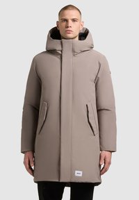 Puffos bézs parka dzseki magas gallérral, nagy kapucnival, két oldalzsebbel és címkével az alsó szélén. Sima anyag, tompa felülettel.