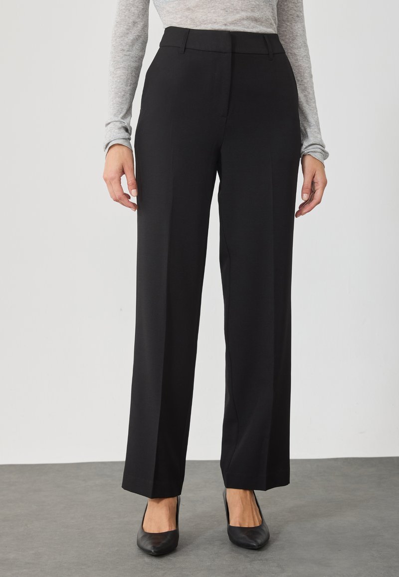 Calças largas pretas com uma textura suave, apresentando uma frente lisa e um corte ajustado, combinadas com sapatos de salto alto pretos com bico afilado.