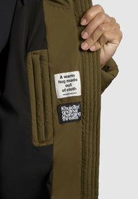 Olivengrøn jakke med sort foer, der har to mærker: et hvidt med tekst og et sort med hvid tekst.