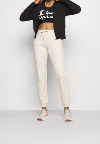 Felpa nera con zip, pantaloni jogger beige con logo bianco e scarpe sportive multicolori. Tessuto liscio e design affusolato con polsini elasticizzati.