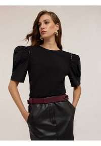 Naisterega, kellel on lainelised pruunid juuksed, on seljas must puff-sleeve top, kuldsed rõngaskõrvarõngad, must nahkne seelik ja tume burdeosvööt, käed taskutes.