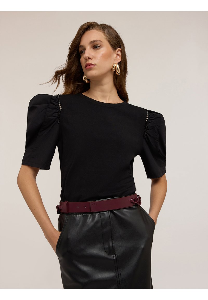 Naisterega, kellel on lainelised pruunid juuksed, on seljas must puff-sleeve top, kuldsed rõngaskõrvarõngad, must nahkne seelik ja tume burdeosvööt, käed taskutes.