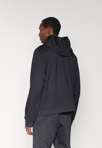 Chaqueta con capucha negra hecha de tela suave, con un ajuste relajado, mangas largas y un diseño minimalista sin logotipos ni patrones visibles.