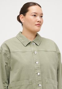 Light green denim jacket featuring a pointed collar, silver snap buttons, and front pockets. Smooth texture with a slightly oversized fit.