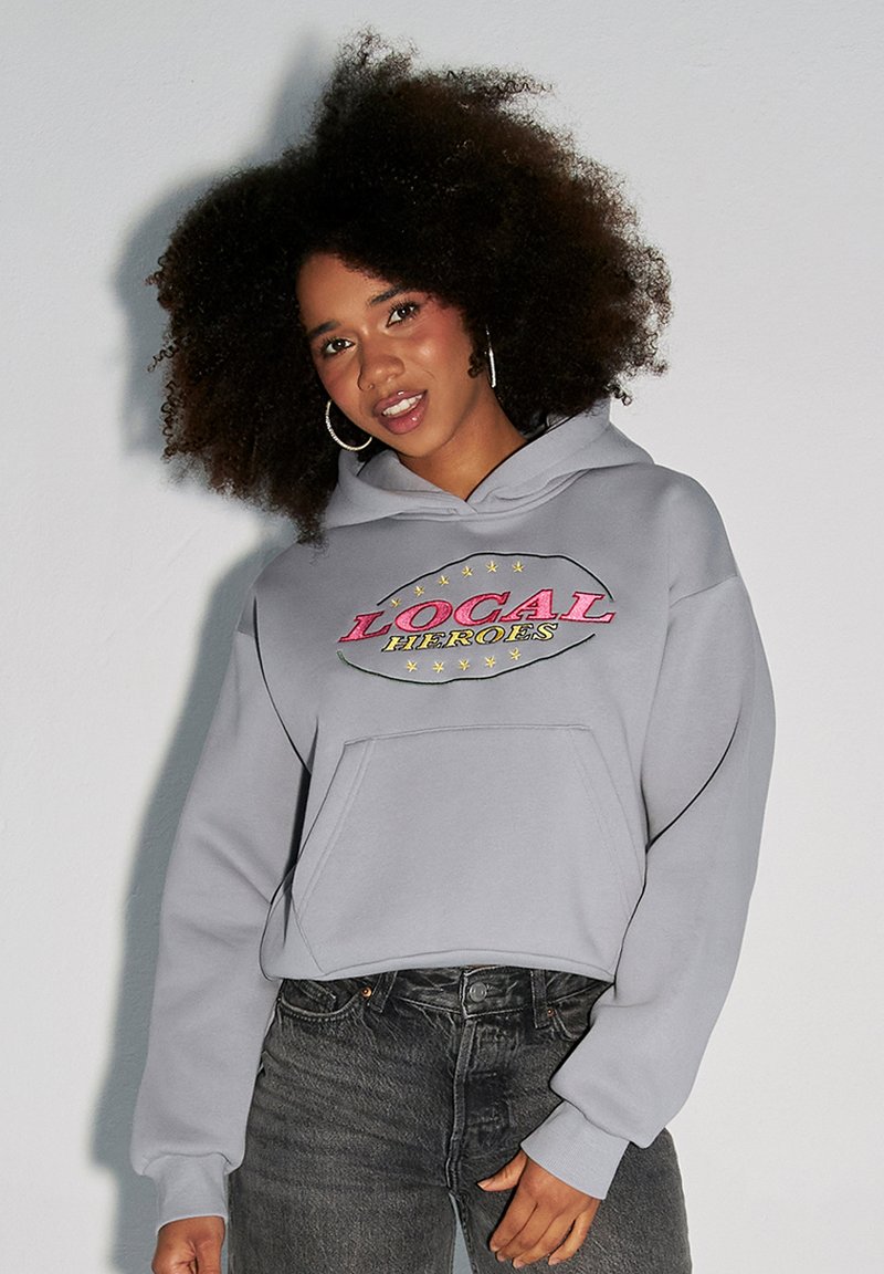 Szary hoodie z kolorową grafiką "LOCAL HEROES", kieszonką z przodu i ściągaczami na rękawach, połączony z ciemnymi dżinsami. Miękki materiał, casualowy fason.
