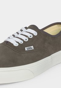 Καφέ αθλητικό Vans με λευκά κορδόνια, λευκή λαστιχένια σόλα και εμφανή ετικέτα με το λογότυπο Vans στο πλάι, σε απλό φόντο.