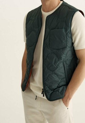 Mężczyzna ubrany w pikowaną, ciemnozieloną kamizelkę bez rękawów na białą koszulę z krótkim rękawem i beżowe spodnie, z rękami w kieszeniach.