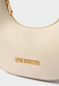 Kremna torba iz gladkega sintetičnega materiala, s zakrivljeno obliko, z zlato tonirano logotipom in zadrgo s čarom v obliki srca.