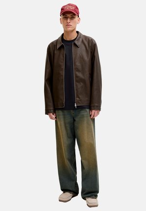 Junger Mann mit roter Mütze, brauner Lederjacke, schwarzem Hemd, ausgeblichenen, lockeren Jeans und beigen Freizeitschuhen, stehend vor weißem Hintergrund.