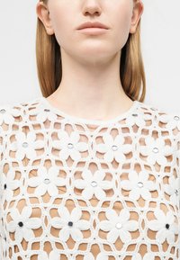 Gros plan d'une femme portant un haut en crochet blanc avec de grands motifs floraux et de petites décorations circulaires réfléchissantes.