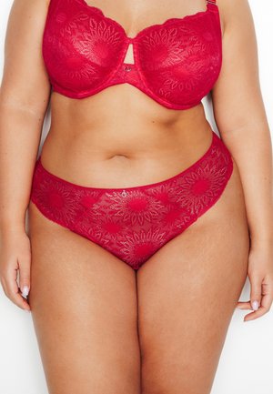 Femme grande taille portant un soutien-gorge en dentelle à fleurs rouges et une culotte assortie, debout les mains détendues le long du corps sur fond blanc.