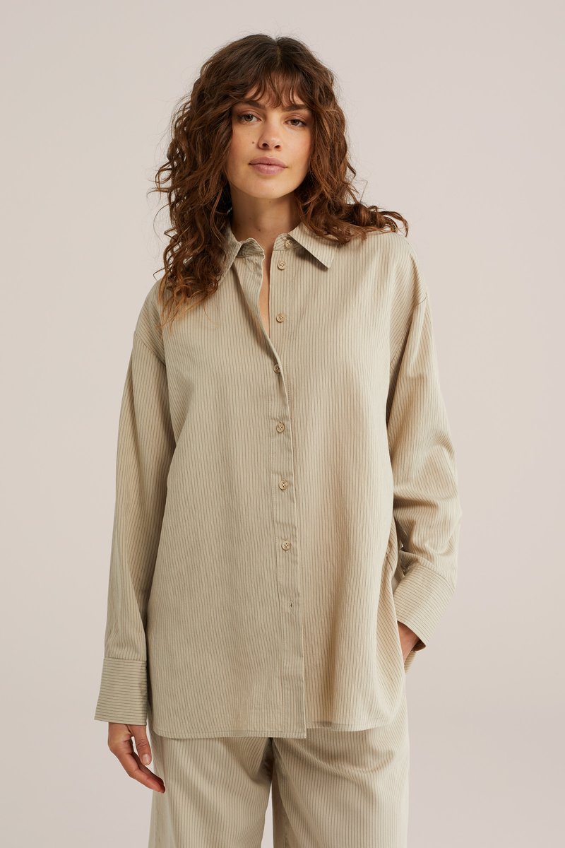 Femme aux cheveux bouclés portant une chemise beige ample à manches longues boutonnée et un pantalon assorti sur un fond uni.