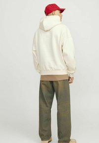 Hvid hætte sweatshirt, brun undertrøje og løse grøna-gule bukser. Personen har en rød kasket på og vender ryggen til kameraet.