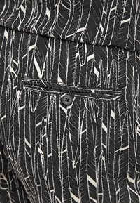 Gros plan d’un pantalon en tissu noir avec un motif de plumes blanches et une poche arrière boutonnée.