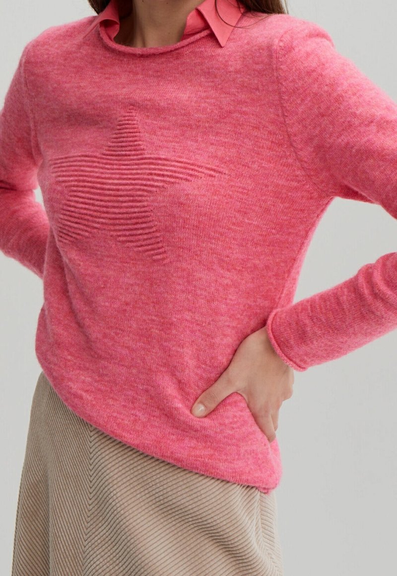 Femme portant un pull rose avec un motif étoile texturé sur une chemise col corail, jupe beige côtelée, main sur la hanche.