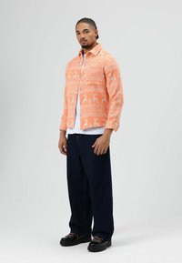 Oranje overhemd met patroon van rendieren en bomen, dichtgeknoopt, over een wit T-shirt. Donkere denim broek met wijde pijpen en zwarte schoenen maken de outfit compleet.