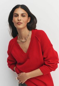 Red knit sweater with a V-neck, ribbed cuffs, and textured sleeves. Model wearing a chain necklace, posing against a white background.