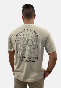 Beige Baumwoll-T-Shirt mit einem bedruckten schwarzen Grafikdesign, das eine Sonne, Strahlen und Text auf der Rückseite zeigt, rundem Halsausschnitt und kurzen Ärmeln.