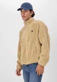 Chaqueta de forro polar beige con un cuello alto, cierre de cremallera, bolsillos laterales y un pequeño parche con un logotipo en el pecho. Llevada con una gorra azul y jeans azules.