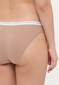 Beige színű pamut hipster alsónemű, fehér elasztikus derékpánttal, amelyen a Lacoste logó található. Simán tapintású, varrás nélküli dizájnnal.