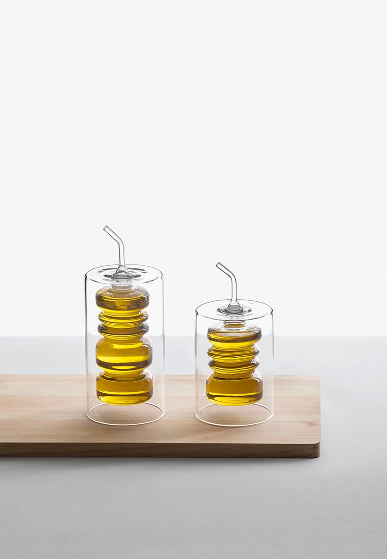 Dwa szklane dozowniki do oleju z warstwowymi, zaokrąglonymi podstawami; jeden wysoki z cienką słomką, drugi krótszy. Oba zawierają złocisto-żółty płyn; wyeksponowane na drewnianej desce.