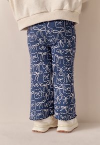 Pantaloni in velluto a coste blu con motivo di fiocchi e nastri bianchi su tutta la superficie, con orli svasati e abbinati a sneakers bianche.