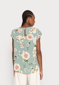 Blusa verde floral com mangas curtas, apresentando flores laranjas e brancas, um detalhe de abertura em forma de buraco nas costas e uma linha de hem do tipo curva.