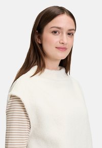 Biały, bezrękawnik o fakturze przypominającej polar, z wysokim kołnierzem. Noszony na jasnej, paskowanej koszuli z długim rękawem. Miękki materiał i minimalistyczny design.