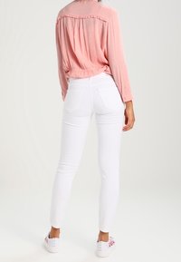 Los vaqueros pitillo blancos tienen una cintura alta y bolsillos traseros, combinados con una camiseta de manga larga en rosa pastel con una costura fruncida en la parte trasera.