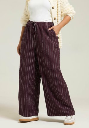 Woman wearing wide-leg dark purple pants with white vertical stripes, white sneakers, white top, and cream textured cardigan with buttons.