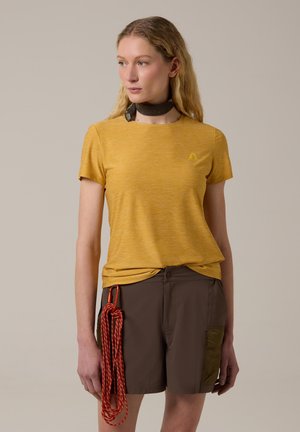 Femme portant une chemise jaune moutarde à manches courtes et un short marron, avec un foulard noir autour du cou et une corde d'escalade rouge enroulée attachée au short.