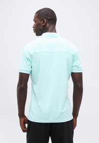 Άνδρας που κοιτάζει προς τα πίσω, φοράει γαλάζιο κοντομάνικο πόλο και μαύρο παντελόνι, μπροστά σε απλό ανοιχτόχρωμο φόντο.