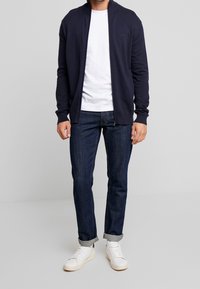 Marinblå zipjacka i bomullsblandning, kombinerad med en vit t-shirt, mörka jeans och vita sneakers. Minimalistisk design och av casual passform.