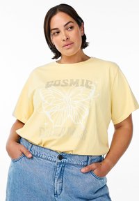 Camiseta de algodón amarilla de corte holgado, con un gráfico de mariposa y acentos de rhinestones, y el texto "Soñador Cósmico" abajo.