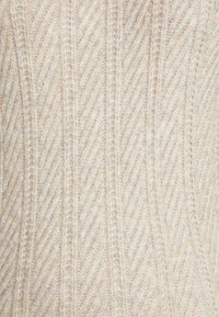 Světle béžový svetr s vzorem herringbone, vyrobený z měkkého, strukturovaného materiálu s jemným leskem.