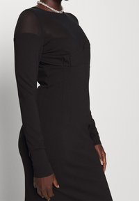 Robe noire avec manches longues, section supérieure en maille transparente, corsage ajusté et texture lisse avec des détails de couture minimaux.