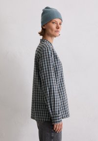 Long-sleeve checkered shirt in blue and black, featuring a buttoned cuff and a rounded collar. Worn with a light blue ribbed beanie.