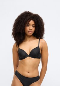 Melnas polsterētas krūšturis ar regulējamām lencītēm, gludu faktūru, smalku spīdumu un dekoratīvu metāla detaļu centrā.